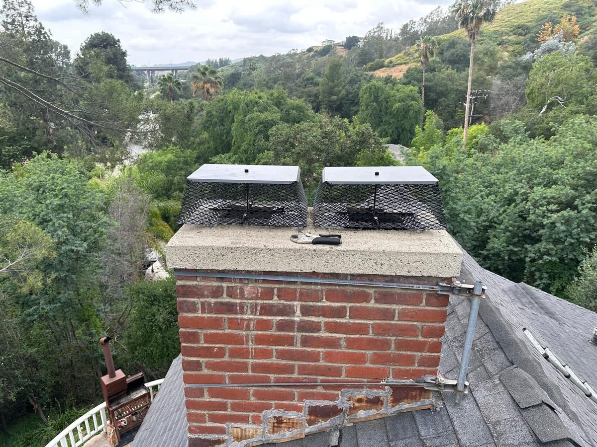 Two newly fitted stainless chimney caps with spark arrestor mesh on a brick chimney above a hillside
