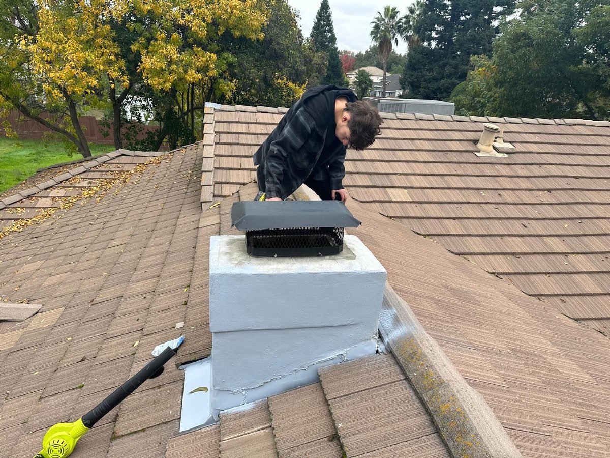 Technician fitting a cap to a freshly sealed chimney crown on a shingle roof