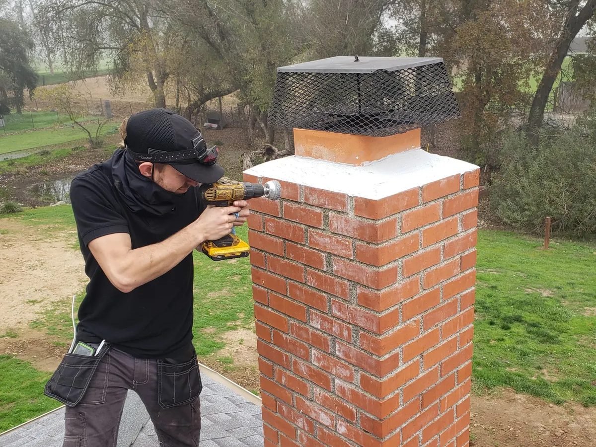 Technician sealing a chimney crown and fitting a new rain cap on a rooftop before the rainy season